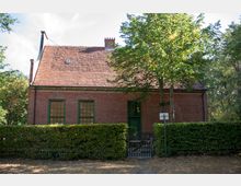 Backsteingebäude mit rotem Ziegeldach inmitten einer grünen, baumreichen Umgebung. Vor dem Haus befindet sich eine niedrige Hecke mit einem Holztor, das den Eingang markiert.