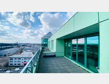 Terrasse eines modernen Gebäudes mit glasverkleideten Wänden und grünlicher Fassade, Blick auf ein Industriegebiet mit flachen Gebäuden und parkenden Fahrzeugen im Hintergrund, unter einem teils bewölkten Himmel.