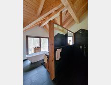 Modernes Badezimmer mit freistehender Badewanne und offener Regendusche. Der Raum hat eine Holzdecke mit sichtbaren Balken, große Fenster mit Blick nach draußen und dunkle Steinfliesen.