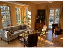 Helles Wohnzimmer mit hohen Sprossenfenstern und einer verglasten Terrassentür, die viel Sonnenlicht auf den Fischgrätparkettboden fallen lässt. Im Raum stehen ein graues Ecksofa, zwei Glas-Couchtische, mehrere klassische Sessel sowie eine Kommode mit großem gerahmtem Porträt und Vorhängen an der Fensterfront.