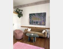 Wohnzimmer mit einem gepolsterten Sofa in Beige, einem rosafarbenen Sessel und einem grünen Marmortisch auf Holzboden. An der Wand hängt eine künstlerische Wanddecke mit abstrakten Motiven, daneben steht eine Monstera-Pflanze; Stuckverzierungen schmücken die Decke.