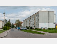 Wohngebiet mit mehrstöckigen, grauen Wohnhäusern im Plattenbaustil. Im Vordergrund befinden sich parkende Autos, eine Straße mit Gehwegen sowie Grünflächen; im Hintergrund steht ein hoher Funkturm.