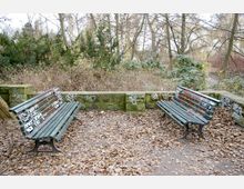 Zwei grüne Parkbänke mit Graffiti stehen sich auf einem mit herbstlichem Laub bedeckten Weg gegenüber, eingerahmt von einer niedrigen Steinmauer und Bäumen im Hintergrund.