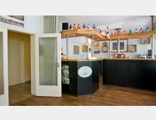 Empfangsbereich eines Hotels mit einer hölzernen Rezeptionstheke, kleinen Dekorationsregalen an der weiß getäfelten Wand und einer Reihe von Länderflaggen auf dem Holzdach der Theke. Links ist eine geöffnete Glastür zu sehen, daneben ein Prospektständer mit Broschüren.