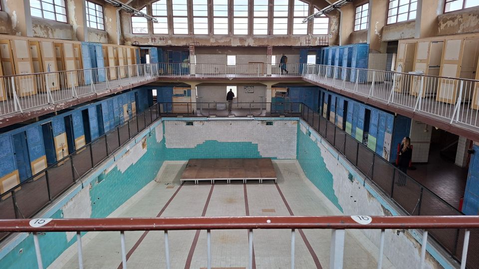 Ein leeres, verfallenes Schwimmbad mit türkisfarbenen Fliesen und verblassten Wänden. Auf zwei Ebenen befinden sich Umkleidekabinen mit blauen und gelben Türen sowie eine Galerie mit Metallgeländern. Große Fenster lassen Tageslicht in den Raum.