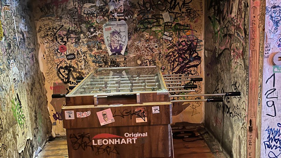 Kleiner, schmaler Innenraum mit Holzboden und niedriger Decke, deren Wände dicht mit Graffiti, Stickern und Kritzeleien bedeckt sind. In der Mitte steht ein Kickertisch (Original Leonhart) unter einer Deckenlampe.