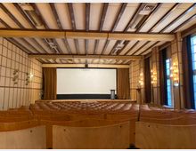 Holzvertäfelter Saal mit einer Bühne im vorderen Bereich, auf der ein Rednerpult und ein geschlossener Vorhang zu sehen sind; davor Sitzreihen aus hellem Holz. An den Seitenwänden befinden sich Wandleuchten und rechts große Fenster mit langen Vorhängen.