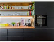 Moderne Küche mit schwarzen Schränken und Holzarbeitsplatte. Offene Regale an der Wand enthalten Bücher, Gläser und Flaschen, während die Arbeitsfläche mit Küchenutensilien, einem Mixer, Schneidebrettern und Dekorationen ausgestattet ist; daneben ein eingebauter Backofen.