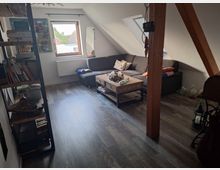 Wohnzimmer mit Dachschrägen, dunklem Holzboden und einer grauen Eckcouch. Ein Holztisch steht in der Mitte, daneben ein Bücherregal und ein Fenster, das Tageslicht hereinlässt.