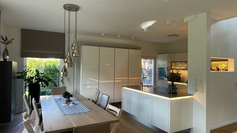 Moderne Wohnküche mit offenem Grundriss, holzfarbenem Esstisch und beige gepolsterten Stühlen. Die Küche verfügt über weiße Hochglanz-Schrankfronten, eine beleuchtete Kücheninsel und eine Glastür mit Blick ins Freie; Pflanzen und Dekorationen ergänzen den Raum.