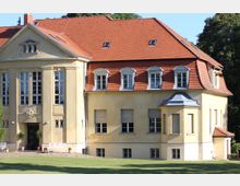 Gelbes, zweigeschossiges Gebäude mit roten Ziegeldächern, großen Fenstern und ornamentalen Details, umgeben von Rasen und Bäumen. Das Bauwerk hat eine symmetrische Fassade mit einem kleinen Erker und mehreren Dachgauben.