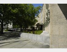 Gebäudefassade aus hellem Stein in einem Kurvenverlauf mit großen Fenstern, davor ein schmaler Grünstreifen mit kleinen Büschen. Links stehen schattenspendende Bäume entlang eines Gehwegs, dahinter sind geparkte Autos und weitere Gebäude sichtbar.