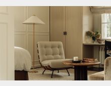 Gemütliches Wohnzimmer oder Schlafzimmer mit einer beigen Sessel-Liege auf einem Teppich, einem runden Holztisch mit einer Kerze und Büchern, einem hohen Stehlampenschirm sowie einem eingebauten Schreibtisch mit Blumen und einer Fensterfront. Die Wände und Möbel sind in hellen, natürlichen Tönen gehalten.