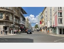 Straßenszene in Berlin mit Blick auf den Checkpoint Charlie, flankiert von historischen und modernen Gebäuden, darunter das „Haus am Checkpoint Charlie“. Einige Passanten stehen an den Gehwegen, während Autos und ein Fahrrad die Kreuzung überqueren.