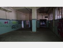 Verlassener Innenraum eines Gebäudes mit kahlen Wänden, abgeblätterter Farbe und türkisfarbenen Fliesen bis zur halben Wandhöhe. Graffiti bedecken die Wände, und durch geöffnete Türrahmen sind weitere Räume sichtbar; die Decke zeigt sichtbare Rohre und Beschädigungen.