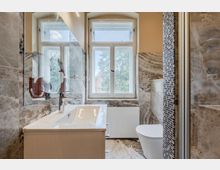 Modernes Badezimmer mit großformatigen marmorierten Wand- und Bodenfliesen, Waschtisch mit Spiegel sowie Toilette neben einer Duschabtrennung mit schmalem Mosaikstreifen. Zwei hohe Fenster über einem Heizkörper lassen Tageslicht herein und bieten Blick ins Grüne.