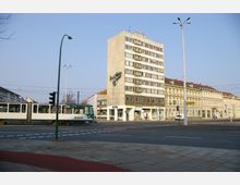 Straßenszene mit einer Straßenbahn, die eine Kreuzung überquert. Im Hintergrund befindet sich ein mehrstöckiges Gebäude mit einer Betonfassade und einer großen Skulptur, daneben historische Gebäude mit hellen Fassaden.