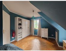 Zimmer mit blauen Wänden und einer Dachschräge, ausgestattet mit einem weißen Kleiderschrank, einem Bücherregal, einer Kommode und einem Fenster mit cremefarbenen Vorhängen. Der Boden ist aus hellem Parkett in Fischgrätenmuster, und ein Feuerlöscher ist an der Wand montiert.