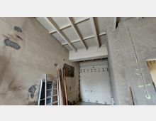 Innenraum einer Baustelle mit unverputzten Wänden, sichtbaren Ziegeln und Betonbalkendecke. An der Wand lehnen Bretter und eine Leiter; der Boden ist uneben, und es sind Arbeiten an der Wandstruktur erkennbar.