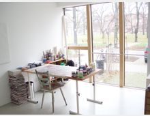 Ein heller Studiobereich mit großen Fenstern, die den Blick auf einen Park mit kahlen Bäumen und einem Gehweg freigeben. Ein Arbeitstisch ist mit Farben und Malutensilien bedeckt, daneben stehen ein Stuhl und ein Stapel Zeitschriften.