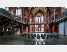 Innenraum einer umgebauten Kirche mit hohen gotischen Spitzbögen, roten Backsteinwänden und großen Fenstern. Der Raum zeigt moderne Elemente wie eine freitragende Metallgalerie und Sitzstühle, während die ursprüngliche Architektur erhalten blieb.