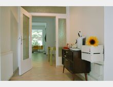 Ein heller Flur mit einer halb geöffneten Glastür, die in einen Raum mit gelben Stühlen und einem großen Fenster führt. Rechts im Flur steht ein Holzschrank mit einer Kaffeemaschine und einer Dekoration in Form einer Sonnenblume.