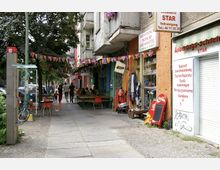 Straßenszene mit kleinen Geschäften und Cafés entlang eines Gehwegs, dekoriert mit bunten Wimpelketten. Im Vordergrund stehen Tische und Stühle unter Bäumen, während Passanten vorbeigehen und Schaufenster mit Waren sichtbar sind.