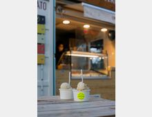 Zwei Becher Eis mit Löffeln stehen auf einem Holztisch im Vordergrund, während im Hintergrund ein beleuchteter Eiswagen mit einer Theke und einer Person sichtbar ist. Die Szenerie wirkt wie ein urbaner Außenbereich.