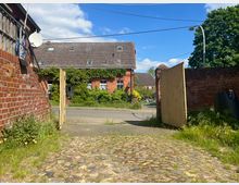 Ein gepflasterter Hof führt durch ein offenes Holztor zu einer ruhigen Straße. Im Hintergrund ist ein Backsteinhaus mit Efeubewuchs und einem grauen Ziegeldach zu sehen, umgeben von Bäumen und einem klaren Himmel.