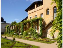 Historisches Gebäude mit ockerfarbener Fassade und verzierten Rundbogenfenstern, flankiert von einer Außentreppe. Im Vordergrund ein gepflegter Garten mit symmetrischen Beeten, Rasenflächen, Blumen und rankendem Grün entlang eines Säulengangs.
