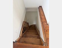 Blick von oben in ein schmales Treppenhaus mit steiler, hölzerner Treppe und Geländer aus Holz. Weiße Wände und ein Holzfußbodenrahmen um die Treppenöffnung prägen den Innenraum.