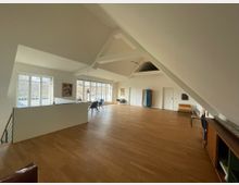 Großzügiger, moderner Wohnraum mit einer Galerie auf der rechten Seite, Holzfußboden und schrägen weißen Deckenbalken. Große Fenster lassen viel Tageslicht herein, und vereinzelte Möbel sowie Kunstwerke an den Wänden akzentuieren den Raum.