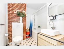 Modernes Badezimmer mit einer markanten roten Ziegelwand, hängender Toilette, weißer Badewanne mit Duschvorhang in Blauverlauf, einem Holzwaschtisch mit Waschbecken und Spiegel sowie schwarz-weiß gemusterten Bodenfliesen. Ein dekorativer Korb und eine weiße Vase mit Blumen sind als Akzente platziert.