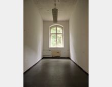 Ein leerer, schmaler Raum mit weißen Wänden und einem rechteckigen Fenster mit Rundbogen, das den Blick auf grüne Bäume nach draußen freigibt. An der Decke hängt eine längliche Deckenleuchte, und der Boden ist dunkelgrau.