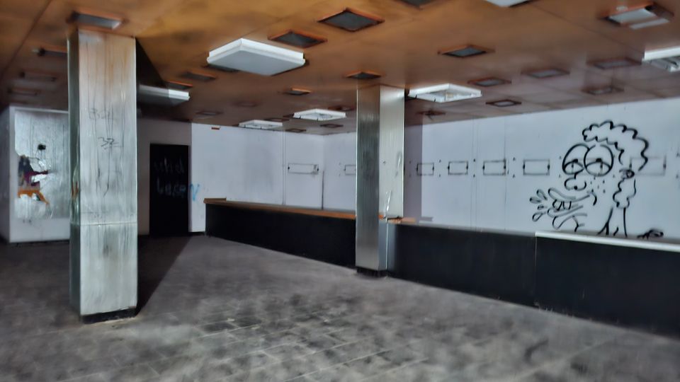 Leerer Innenraum eines verlassenen Laden- oder Gastronomiebereichs mit langer Theke entlang der Rückwand, Metallstützen und abgehängter Decke mit rechteckigen Leuchten. Weiße Wandpaneele mit kleinen Öffnungen und Graffiti, dunkler Fliesenboden.