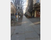 Straßenszene in der Oderberger Straße mit breitem, gepflastertem Gehweg, flankiert von kahlen Bäumen und mehrstöckigen Altbauhäusern. Am rechten Gehwegrand befinden sich Cafés und Geschäfte, links sind Fahrräder geparkt.