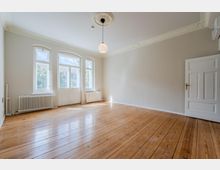 Heller, leerer Altbau-Wohnraum mit hohen Decken, Stuckverzierungen und einem runden Deckenleuchter. Drei hohe Fenster und eine verglaste Flügeltür lassen viel Tageslicht auf den Holzdielenboden; an den Wänden stehen Heizkörper und eine weiße Zimmertür ist rechts sichtbar.