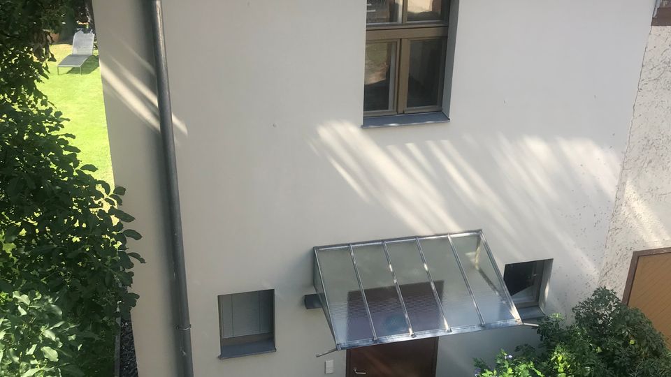 Kleines, zweigeschossiges Gebäude mit heller Fassade und Flachdach, mit einem zentralen Fenster im Obergeschoss und einem Eingang im Erdgeschoss unter einem gläsernen Vordach. Vor dem Eingang führen wenige Stufen hinauf, umgeben von Büschen und Bäumen; im Hintergrund ist ein größeres rotes Backsteingebäude mit vielen Fenstern zu sehen.