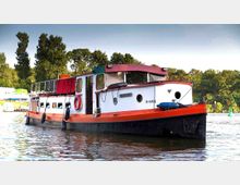 Ein rot-weißes Hausboot mit Flachdeck und mehreren Fenstern liegt auf einem Gewässer, umgeben von grünen Bäumen und anderen Booten im Hintergrund. Am Boot sind Rettungsringe und Fender angebracht, die Kulisse zeigt eine ruhige, bewaldete Uferlandschaft.