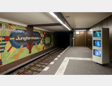 U-Bahnhof Jungfernheide: Ein unterirdischer Bahnsteig mit bunt gestalteten Wandfliesen, die geometrische Muster zeigen. Links befindet sich der Schriftzug "Jungfernheide", während rechts Monitore und eine Uhr sichtbar sind; darüber hinaus führen Schienen in einen dunklen Tunnel.