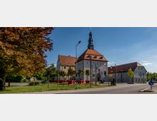 Historisches Schlossgebäude mit hellen Fassaden und rotem Ziegeldach, flankiert von einem schlanken Turm. Im Vordergrund steht „SCHLOSS“ als Installation mit großen Buchstaben, umgeben von Bäumen und grünem Rasen, bei klarem Himmel.