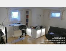 Ein kleiner, heller Arbeitsraum mit einem höhenverstellbaren Schreibtisch, einem weißen Stuhl, einem Regal voller Bücher und einem Sofa. Zwei kleine Fenster mit gemusterten Vorhängen und ein niedriger Schrank mit Beleuchtung ergänzen die Einrichtung.