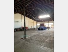 Große Lagerhalle mit sichtbarem Holzdach und Metallstützen, die einen offenen, leeren Innenraum umrahmen. An den Wänden stehen diverse Werkzeuge, Regale und Gerätschaften, während in der Mitte ein mit einer Plane abgedecktes Fahrzeug steht.