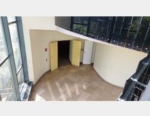 Treppenhaus-Bereich mit hellen Fliesenboden, gelb gestrichenen Türen und großen Fenstern mit Blick auf eine begrünte Außenanlage. Eine gewundene Wand und ein schwarzes Metallgeländer mit einfallendem Sonnenlicht prägen den Raum.