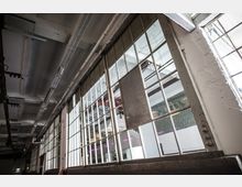 Industrieller Innenraum mit großen, drahtverstärkten Fenstern, die Licht ins Gebäude lassen. Sichtbare Deckenstruktur mit Kabelkanälen und Beleuchtung, sowie weiß verputzte Wände.