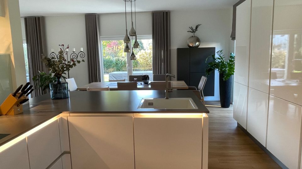 Moderne, offene Küche mit weißen Hochglanzschränken, einer zentralen Kücheninsel mit Spüle, Holzfußboden und großen Fenstern, die viel Tageslicht hineinlassen. Im Hintergrund befindet sich ein Essbereich mit Tisch, Stühlen und großen Zimmerpflanzen.