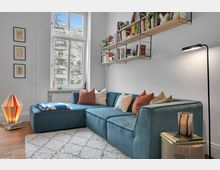 Helles Wohnzimmer mit einem großen blauen Ecksofa, mehreren farbigen Kissen und einem weißen Teppich mit geometrischem Muster auf Holzboden. An der Wand ein großes Fenster, darüber schwebende Bücherregale mit Büchern und Dekorationen; links eine Tischlampe, rechts eine Stehlampe.