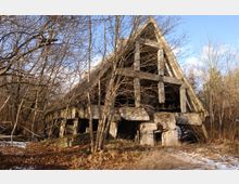 Verfallenes Gebäude mit spitzem Giebel, bestehend aus Beton und von Bäumen und Gestrüpp umgeben. Der Boden ist teilweise mit Laub und Schnee bedeckt, und im Hintergrund ist ein blauer Himmel zu sehen.