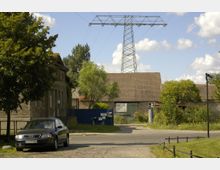 Straßenansicht in einem ländlichen Gebiet mit einem geparkten Auto auf gepflasterter Fahrbahn, einem alten Gebäude mit verputzten Wänden auf der linken Seite und einer Scheune mit Ziegeldach im Hintergrund. Im oberen Teil ragt ein großer Strommast in den Himmel, umgeben von vereinzelten Bäumen und wolkigem Himmel.