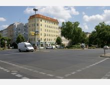 Straßenkreuzung in einer städtischen Umgebung mit mehrstöckigen Wohnhäusern im Hintergrund, darunter ein gelbes Gebäude mit roten Dachziegeln. Rechts sind Grünflächen mit großen Bäumen sichtbar, ein U-Bahn-Schild und Straßenschilder sind ebenfalls vorhanden.